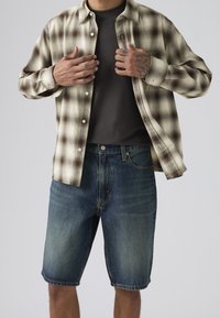 Άνδρας που φοράει ξεθωριασμένο μπλε τζιν σορτς, σκουρό γκρι μπλουζάκι και μπεζ πουκάμισο με καρό μοτίβο, μερικώς ανοιχτό, προσαρμόζοντάς το με τα τατουάζ χέρια του.