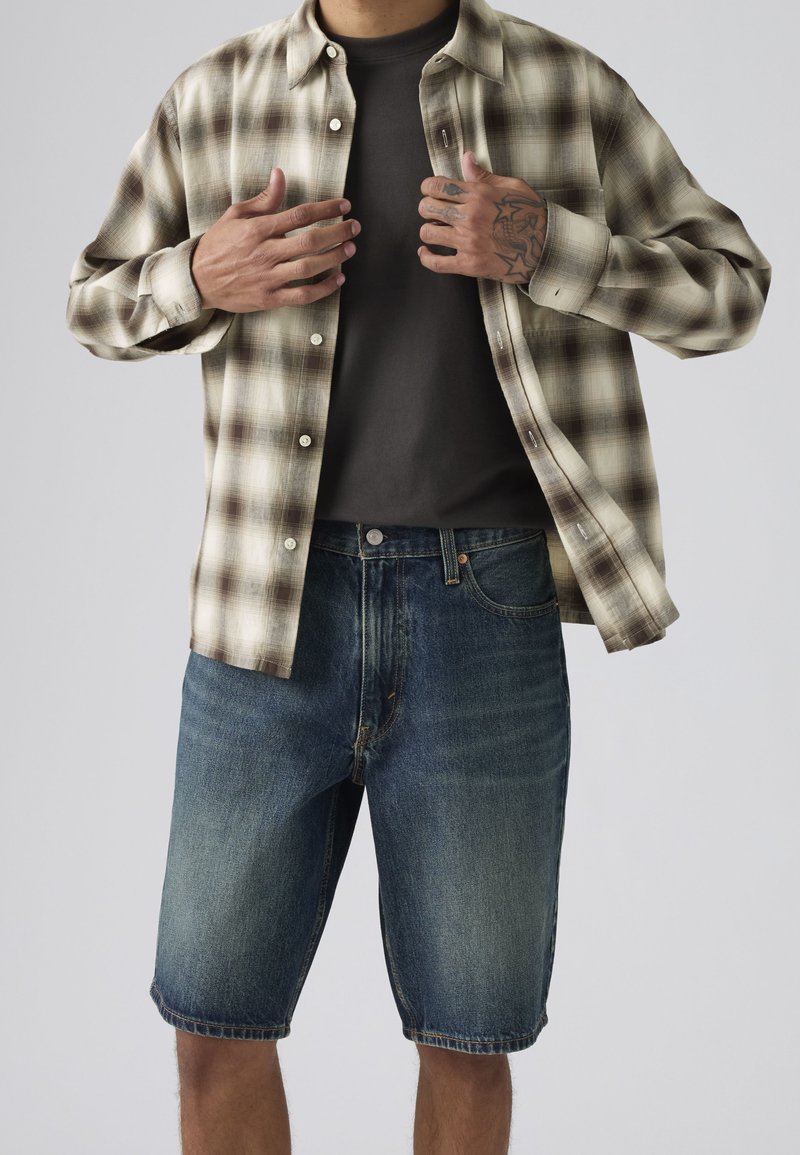 Homme portant un short en jean bleu délavé, un t-shirt gris foncé et une chemise à carreaux beige partiellement ouverte, ajustée par des mains tatouées.