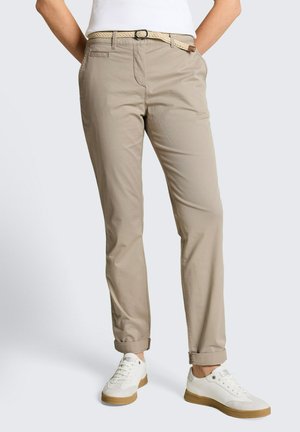 Osoba nosząca beżowe spodnie chino z podwiniętymi mankietami, pleciony beżowy pasek, białe trampki z gumowymi podeszwami i biały bezrękawnik.