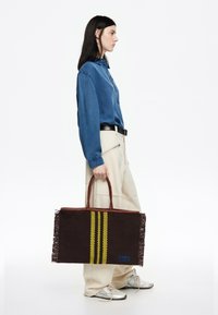 Bruinegeweven tas met franjes, voorzien van twee gele strepen en blauwe branding. Leren handvatten, rechthoekige vorm en gestructureerd ontwerp.