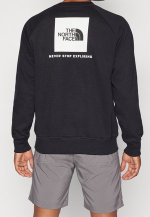 Persoon draagt een zwart sweatshirt met wit "The North Face" logo en "Never Stop Exploring" tekst op de rug, gecombineerd met grijze shorts.