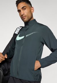 Tummanvihreä Nike vetoketjullinen takki, jossa on korkea kaulus, ja siinä on vaalean teal värinen swoosh-logo sekä joustavat hihansuut. Suunniteltu aktiiviseen käyttöön.