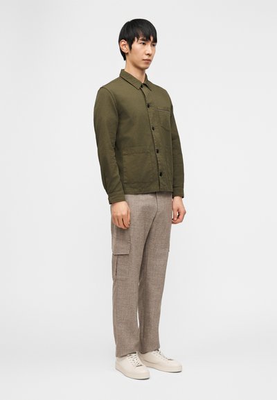 Άνδρας με χακί πράσινο σακάκι με κουμπιά, ανοιχτό καφέ παντελόνι cargo και λευκά αθλητικά παπούτσια, που στέκεται μπροστά σε ένα απλό ανοιχτό φόντο.