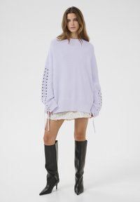 Lila übergroßer Strickpullover mit Rippstruktur und Schnürärmel, kombiniert mit einem weißen Spitzen-Minirock und knielangen schwarzen Stiefeln.