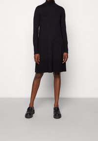 Abito nero a maniche lunghe con design a collo alto. Realizzato in tessuto morbido, presenta una silhouette dritta e sottili aperture laterali. Mocassini neri.