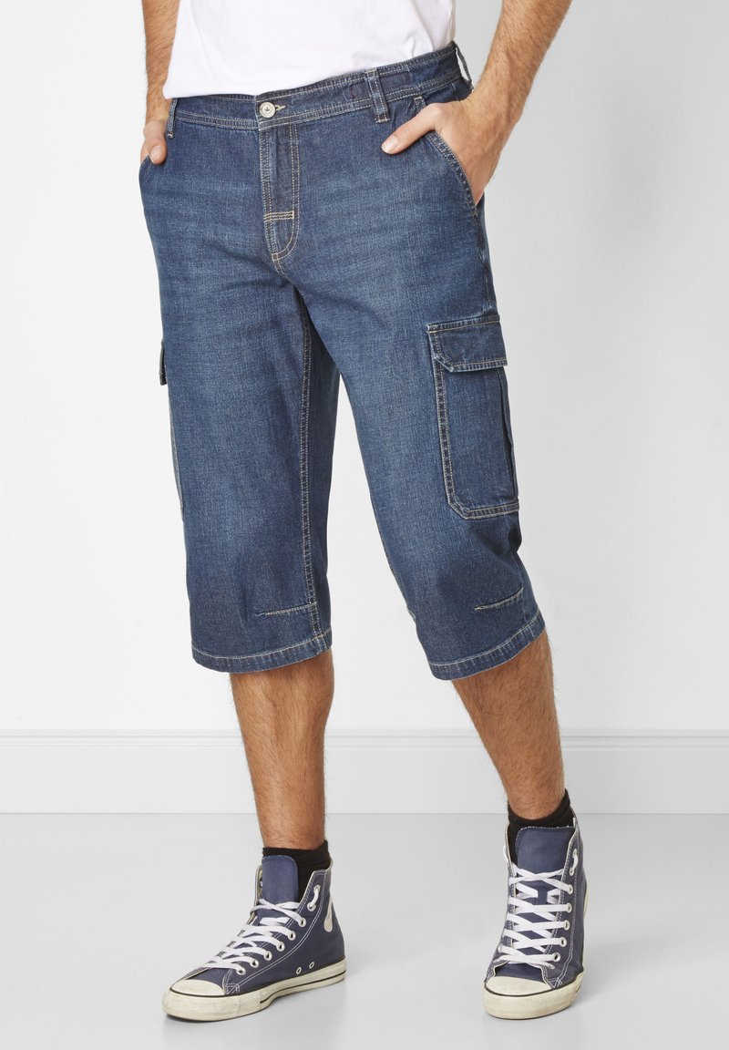 Denim cargo shorts in dark blue with knee-length cut, side pockets, and contrasting stitching details. Paired with high-top sneakers.