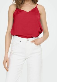 Haut rouge sans manches avec encolure festonnée, en tissu lisse, associé à un jean blanc taille haute avec un détail à boutons à la taille.