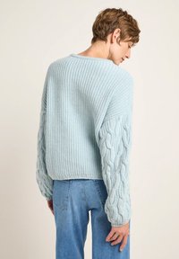 Hellblauer Strickpullover mit strukturiertem Kabeldesign an den Ärmeln, geripptem Muster am Körper und lässiger Passform. Getragen mit blauen Jeans.