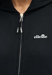Κοντινό πλάνο μαύρου φούτερ με φερμουάρ και λευκό λογότυπο ellesse στο στήθος, με φερμουάρ που φέρουν το εμπορικό σήμα.