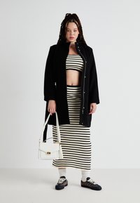 Svart frakk, stripet crop topp og lang skjørt i beige og svart. Hvit håndveske, mønstrede sko og ribbet tekstur. Enkel design.