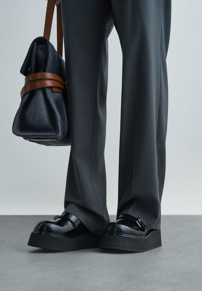 Persoană îmbrăcată cu pantaloni gri croiți și loafers negri cu talpă înaltă, purtând o geantă bleumarin cu mânere din piele maro.