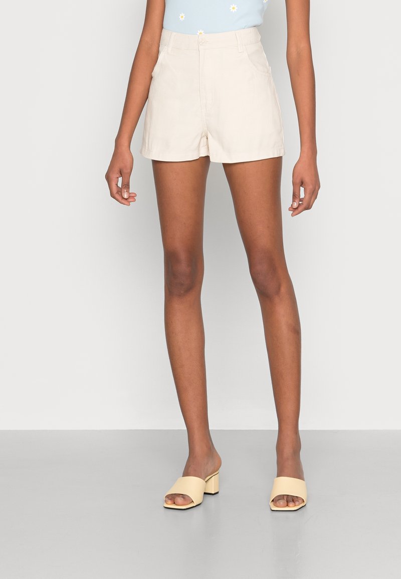 Femme portant un short taille haute beige, un haut bleu clair fleuri et des sandales ouvertes à talons jaunes pâles, debout devant un fond uni.