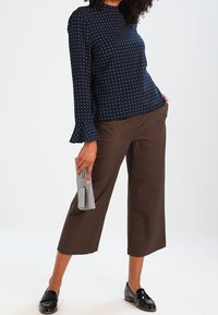 Donna che indossa una blusa a maniche lunghe a pois blu navy, pantaloni cropped larghi marroni, mocassini neri e tiene una pochette grigia in una mano.