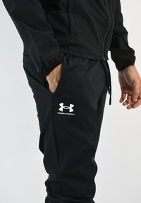 Chaqueta y pantalones deportivos negros hechos de una tela suave, con puños elásticos, bolsillos laterales y un logo blanco de Under Armour en los pantalones.