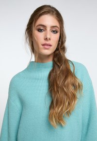 Pulover tricotat de culoare albastru deschis, cu un guler înalt și textură ribbată, având mâneci largi și o croială relaxată. Părul neted cade pe o parte.