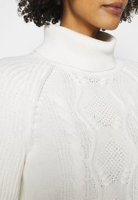 Femme portant un pull à col roulé en tricot blanc avec des motifs torsadés et en losanges, cheveux bouclés visibles au niveau de l'encolure.