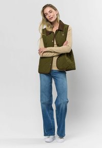 Pikowana oliwkowa kamizelka z brązowym kołnierzem, metalowymi zatrzaskami i luźnym krojem, warstwowa na beżową bluzkę z długim rękawem oraz niebieskie dżinsy.