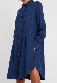 Denimová košilová šaty v tmavě modré, dlouhé rukávy, přední zapínání na knoflíky, boční kapsy a zaoblený spodní lem s volným střihem.