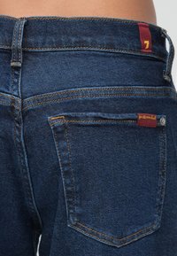 Sötétkék farmernadrág narancssárga varrással, hátsó zsebbel, melyen egy kis piros címke található, valamint derékpánt hurkakkal.