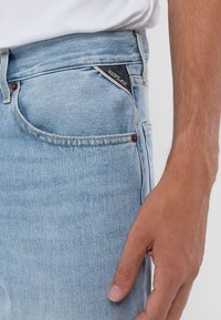 Ljusblå denimjeansficka med en liten mörk triangelmärkning och en persons högra hand vilande bredvid.