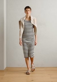 Robe midi à rayures noires et blanches en tissu extensible, associée à un pull beige clair drapé sur les épaules et des sandales marron.
