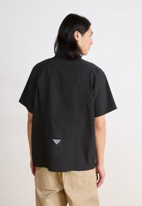 Black short-sleeve shirt with a relaxed fit, featuring a triangular accent at the back and a smooth, lightweight fabric texture.