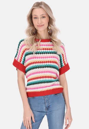 Maglione corto a maniche corte dai colori vivaci con motivo a zigzag nei toni di rosa, verde, rosso e bianco. Collo e orlo rossi a costine. Indossato sopra jeans azzurri chiari.