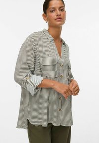 Femme portant une chemise beige et blanche à rayures verticales, à manches longues et à coupe ample, avec deux poches avant, associée à un pantalon vert foncé.