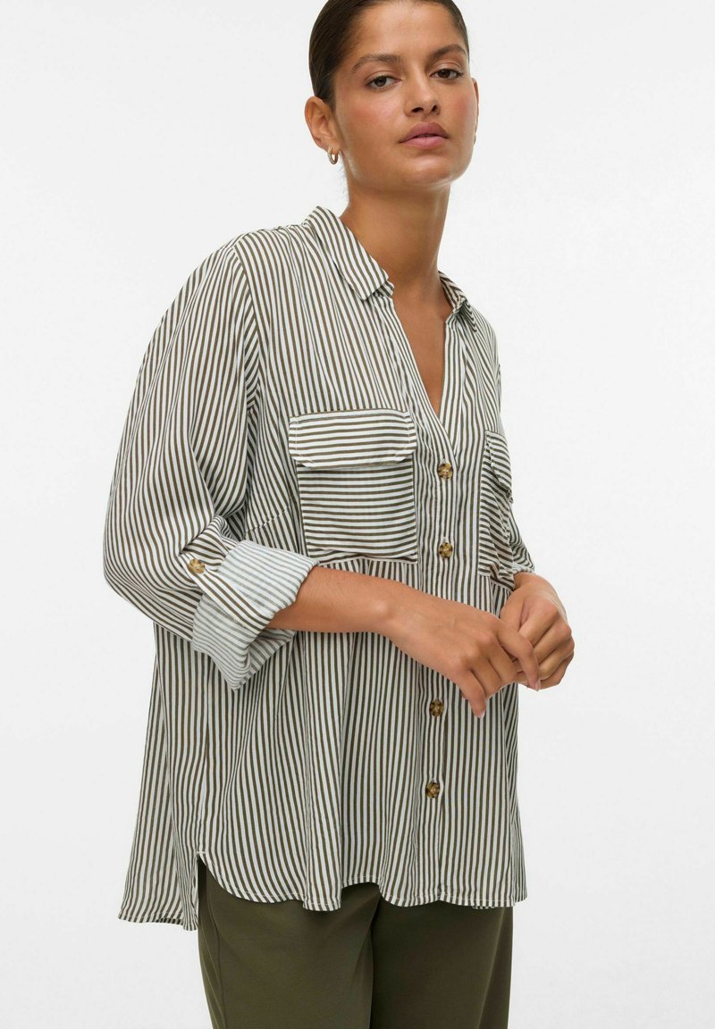 Femme portant une chemise beige et blanche à rayures verticales, à manches longues et à coupe ample, avec deux poches avant, associée à un pantalon vert foncé.