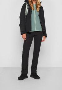 Sort isoleret jakke, grøn zip-up bluse, sorte bukser og sort fodtøj. Har en rygsæk med lette stropper. Simpel, funktionel design.