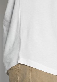 Weißes Baumwoll-T-Shirt mit abgerundetem Saum, das eine glatte Textur und kontrastierende Nähte aufweist, über beigem Stoff geschichtet.