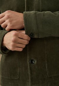 Olive zaļa korduroja krekla jaka ar ribotu tekstūru, melnām pogām un pogājamiem piedurkņu galiem. Redzamas rokas, kas pielāgo piedurkņu detaļu.