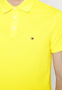 Sárga polo póló texturált anyaggal, háromgombos záródással és egy kis logóval, piros, fehér és kék díszítéssel a bal mellrészen.