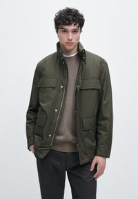 Olivengrøn jakke med høj krave, to forlommer med påsyede klapper og trykknapper. Båret over en lysebrun sweater og mørke bukser.