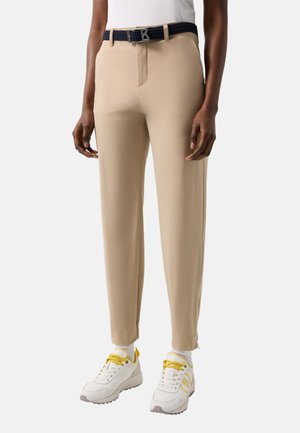 Personne portant un pantalon beige sur mesure, une ceinture noire avec une boucle en forme de "B", des baskets blanches avec des lacets jaunes, et des chaussettes blanches, debout devant un fond blanc.