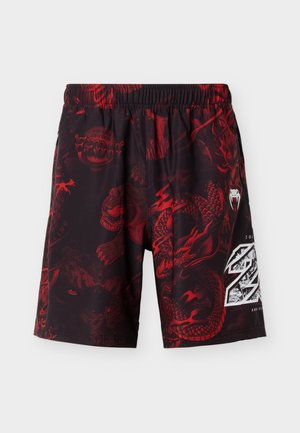 Μαύρα και κόκκινα σορτς με ελαστική μέση, διακοσμημένα με γραφικά δράκου και κρανίου και λευκό έμβλημα επετείου στο δεξί πόδι.