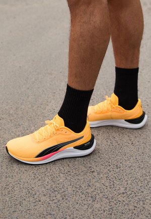 Des chaussures de course jaunes vives avec des accents noirs, un dessus texturé, une semelle blanche et un détail rose au talon. Portées avec des chaussettes noires.