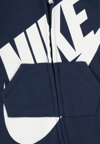 Tengerészkék cipzáros kapucnis pulóver nagy fehér Nike logóval, kenguruzsebbel és kontrasztos varrással. Puha pamut anyagból.