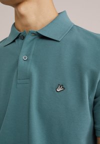 Polo de color verde azulado hecho de tela texturizada, con una tapeta de tres botones y un logo de ave bordado en el lado izquierdo del pecho.