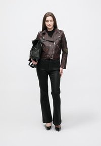 Bordeauxrode leren bikerjack met studs en patches, gecombineerd met zwarte strakke broek en zwarte hoge hakken. Model houdt een zwarte tas vast.