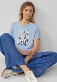 T-shirt léger bleu clair à manches courtes avec un graphique d'architecture grecque blanche et un label "Grèce". Porté avec un pantalon large bleu.