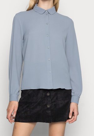 Blouse bleu clair à manches longues avec un boutonnage sur le devant, petit col et poignets à fronces, associée à une jupe en velours côtelé noire dotée de boutons.