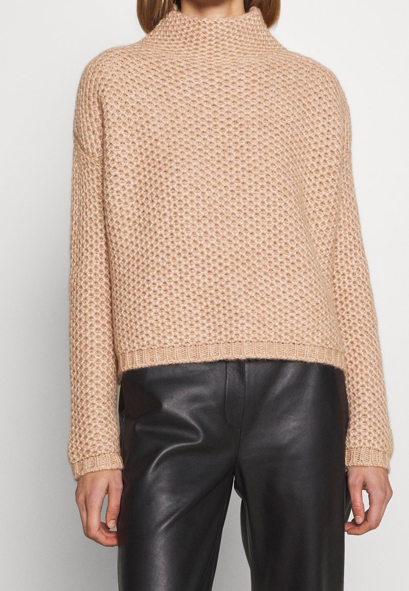 Pull en maille beige texturé avec col montant et manches longues associé à un pantalon en cuir noir sur une personne debout sur fond blanc.