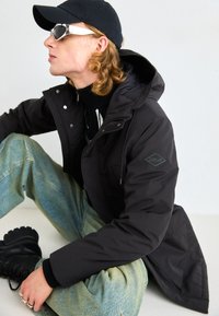 Giacca a vento nera con colletto alto, caratterizzata da una patch con logo, abbinata a jeans azzurri chiari e stivali da combattimento neri. Gli accessori includono occhiali da sole e un cappellino.