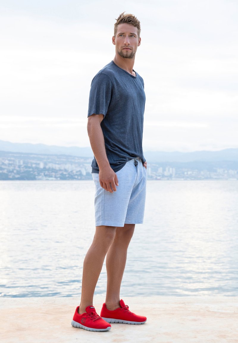 T-shirt gris à manches courtes, shorts bleu clair et chaussures de sport rouges avec une tige texturée. Cadre extérieur avec un arrière-plan aquatique.