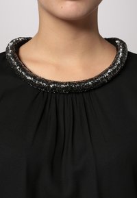 Zwarte blouse met een gathered halslijn en een chunky, metallic zilveren kralencollar. Het gladde materiaal zorgt voor een strakke uitstraling.