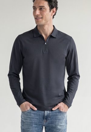 Homme portant un polo noir à manches longues avec une fermeture éclair à l’avant, mains dans les poches, regardant sur le côté sur un fond uni.