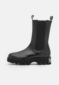 Black leather ankle boots with elastic side panels, rounded toe, thick black sole, and white logo detail on the heel. Matte and shiny finish.