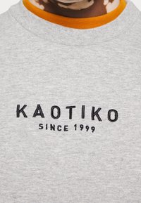 Grijze sweater met ronde hals en oranje binnenkraag. Voorzien van zwart geborduurde tekst "KAOTIKO" en "SINCE 1999" op de voorkant.