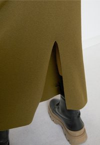 Olijfgroene gebreide jurk met een zijsplit, gecombineerd met zwarte schoenen met een dikke zool, met een gladde textuur en eenvoudig ontwerp.